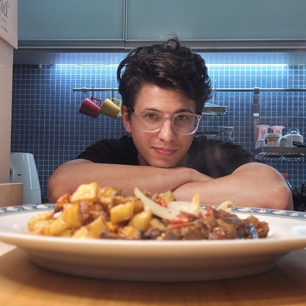 Tobias ao fundo em sua cozinha diante de um prato de comida